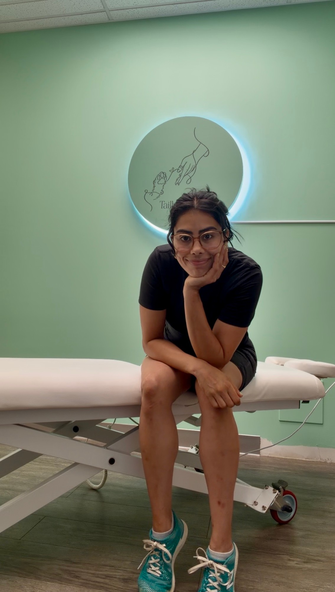 A smiling Tailbone Health practitioner sitting on a treatment table in front of the clinic logo