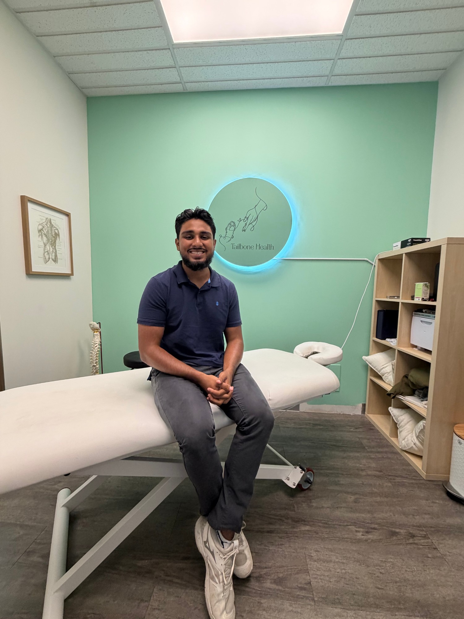 A Tailbone Health practitioner smiling in a treatment room beneath the illuminated logo