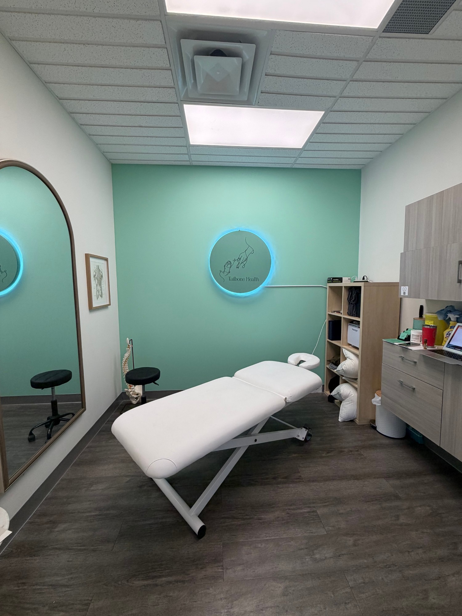 A Tailbone Health treatment room with a mint feature wall, the illuminated clinic logo and a treatment table